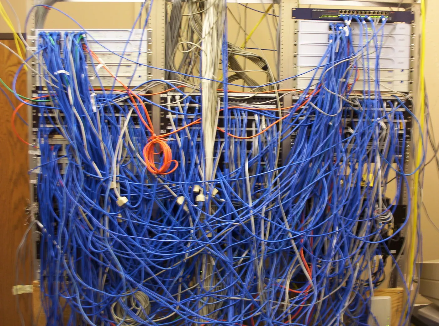 Stock photo: a network closet where a thick tangle of blue and gray cables hangs in front of the equipment.