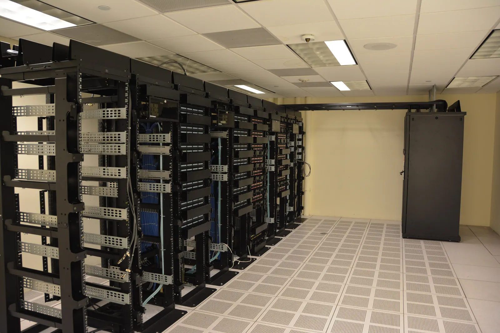Stock photo: a quiet equipment room with rows of open black racks and patch panels under ceiling lights. Nobody is in the room.