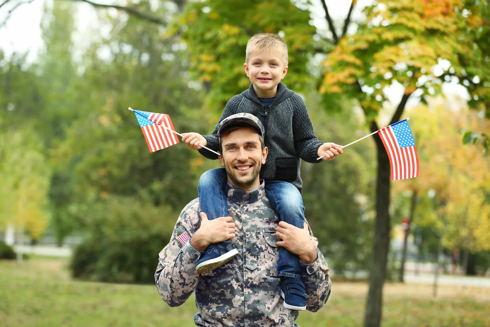 A U.S. servicemember carrying a young boy on his shoulders outdoors, the boy holding a small American flag in each hand