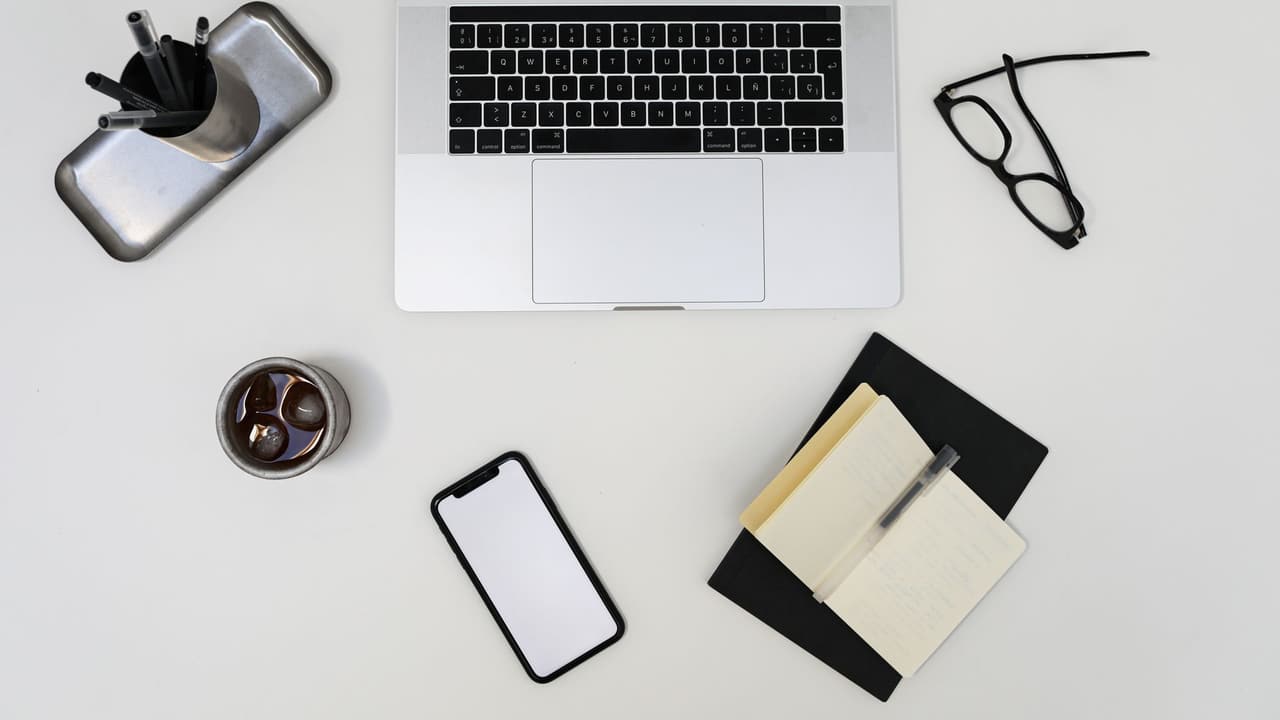 Stock photo: a laptop, a generic smartphone with a blank screen, reading glasses and a notebook seen from above on a light desk.